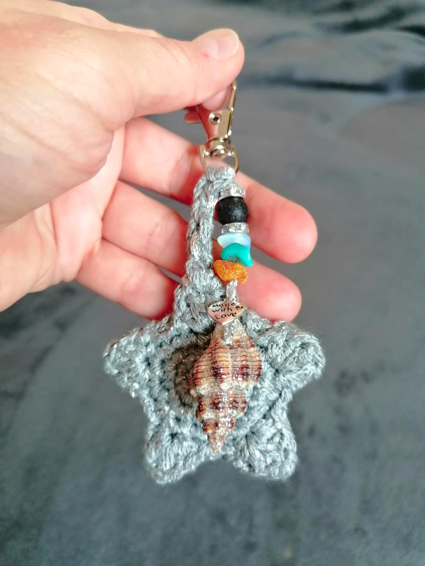 A grey crocheted star keychain with beads and a shell, held in a hand