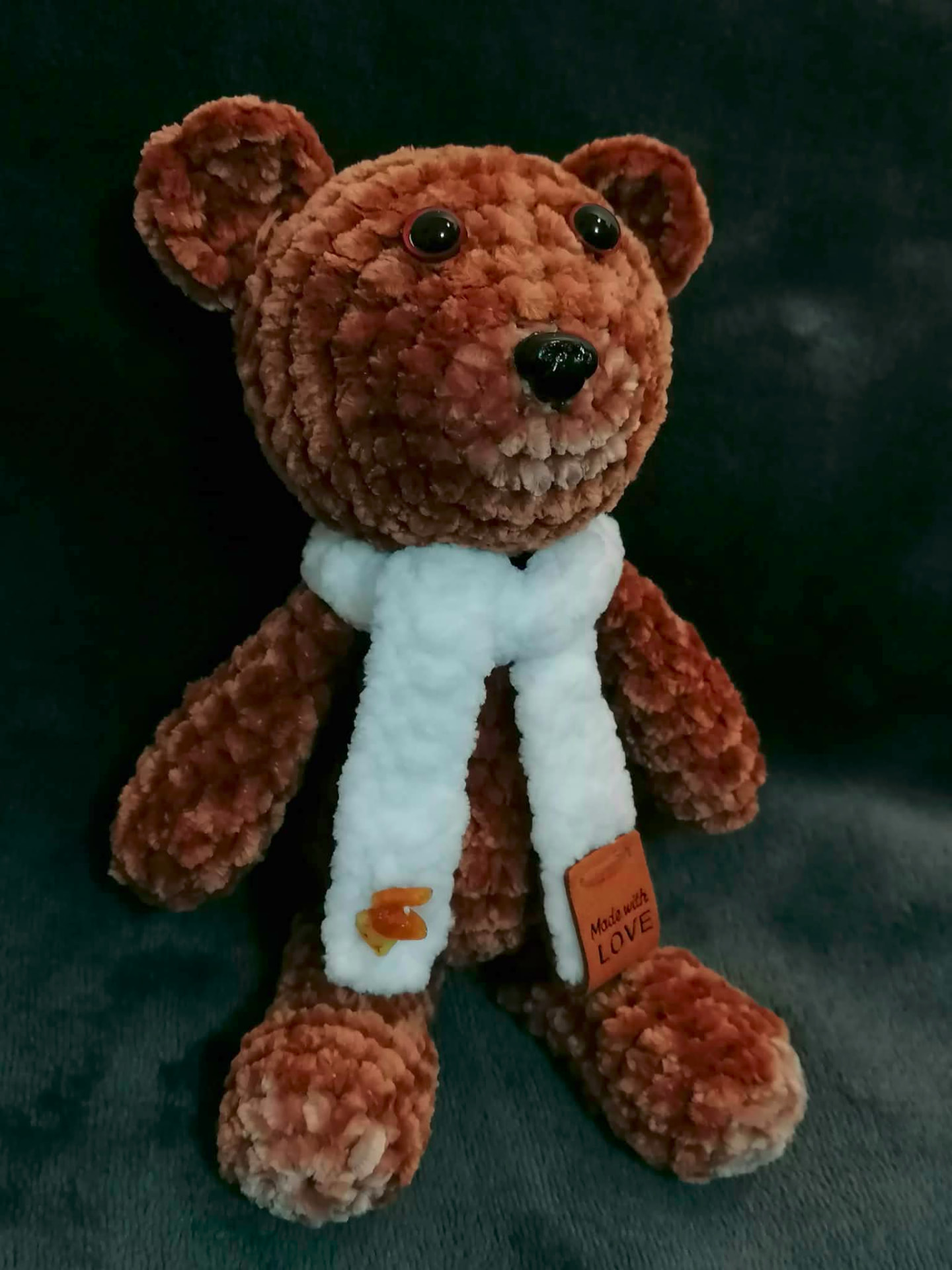 Close-up of a handmade crocheted brown bear wearing a white scarf with a small amber heart bead and a Made with Love tag