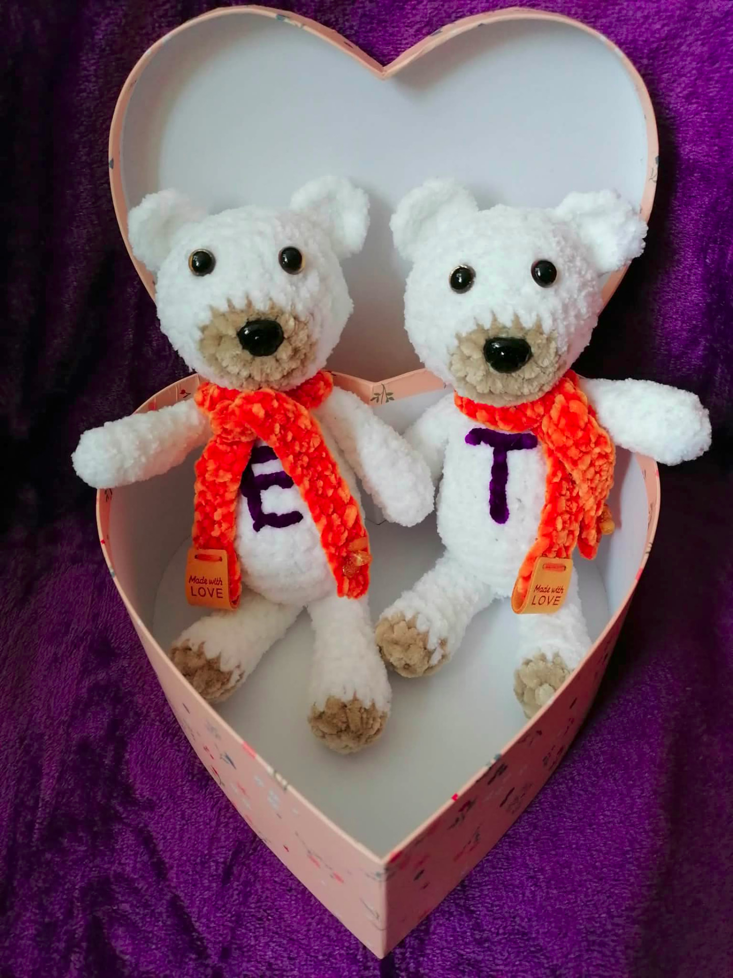Two white handmade crochet teddy bears with embroidered initials and orange scarves, presented in a heart-shaped box