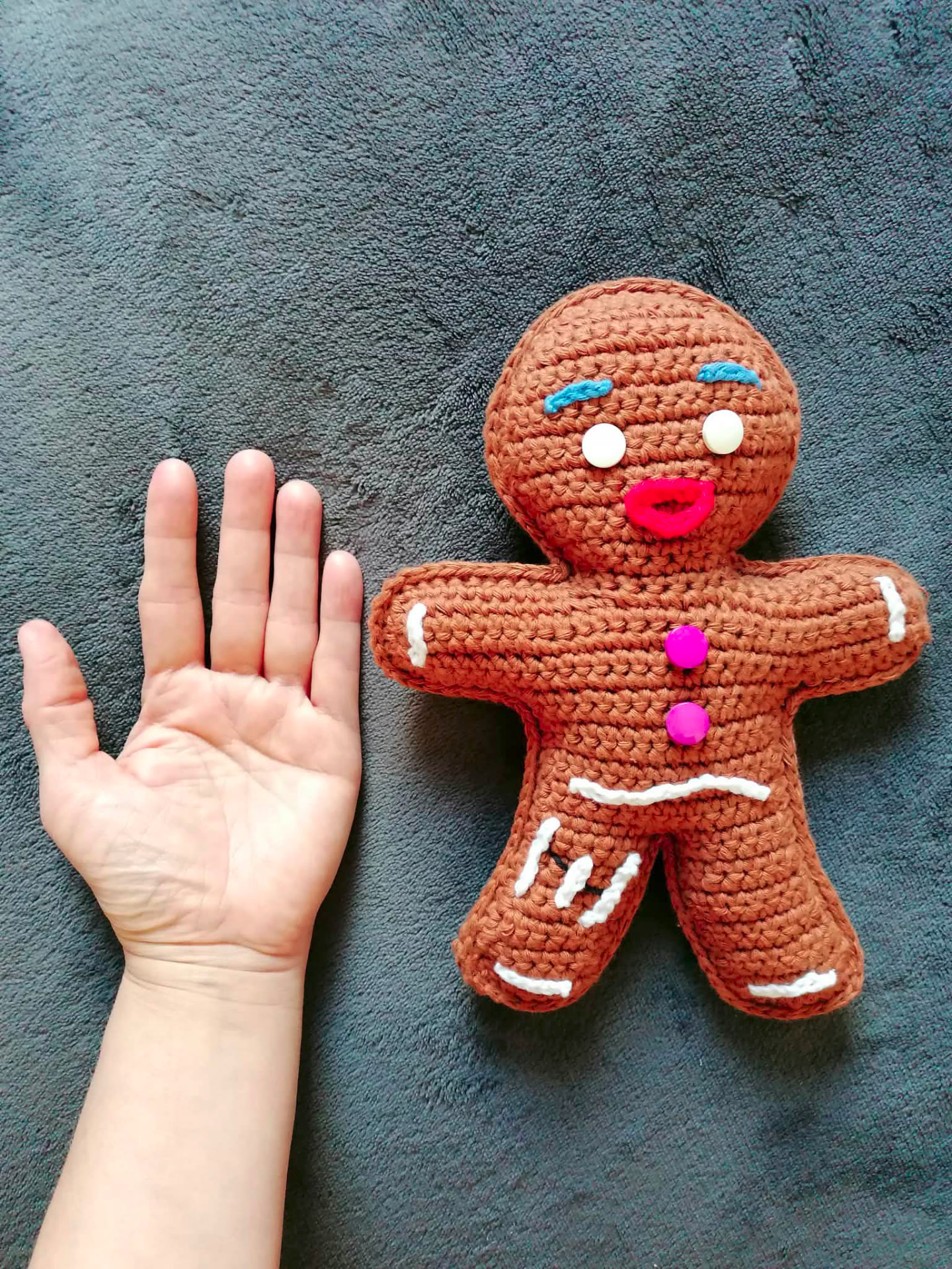 A large handmade crocheted gingerbread character with white icing details, shown beside a hand