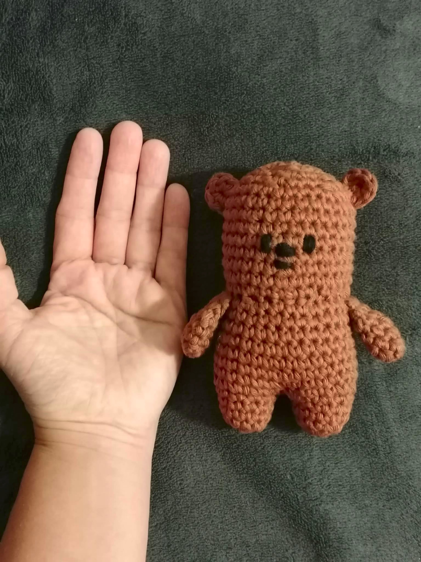 A small handmade crocheted teddy bear in warm brown tones beside a hand