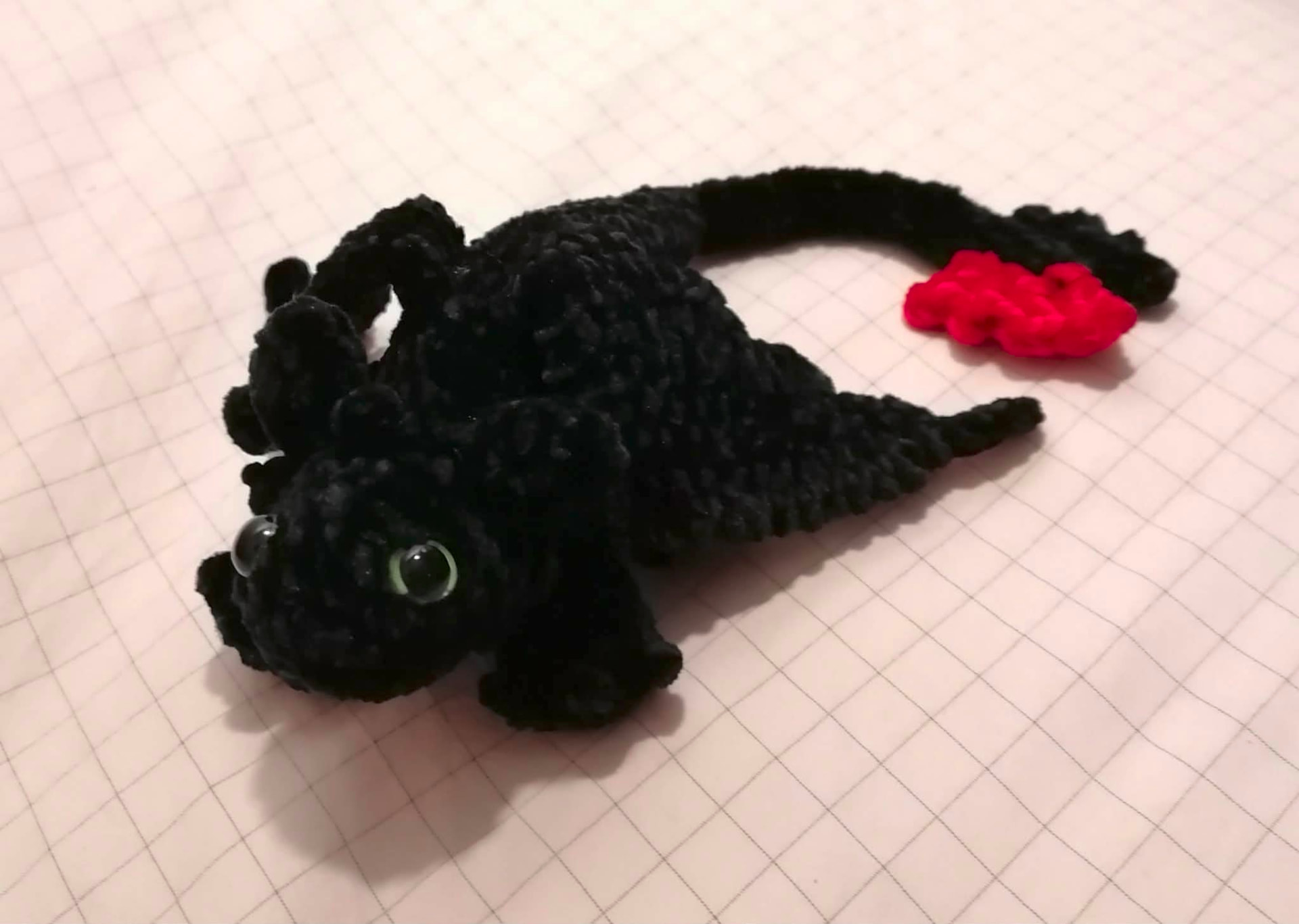 A handmade black crocheted dragon with green eyes and a long tail, shown in full beside a small red flame