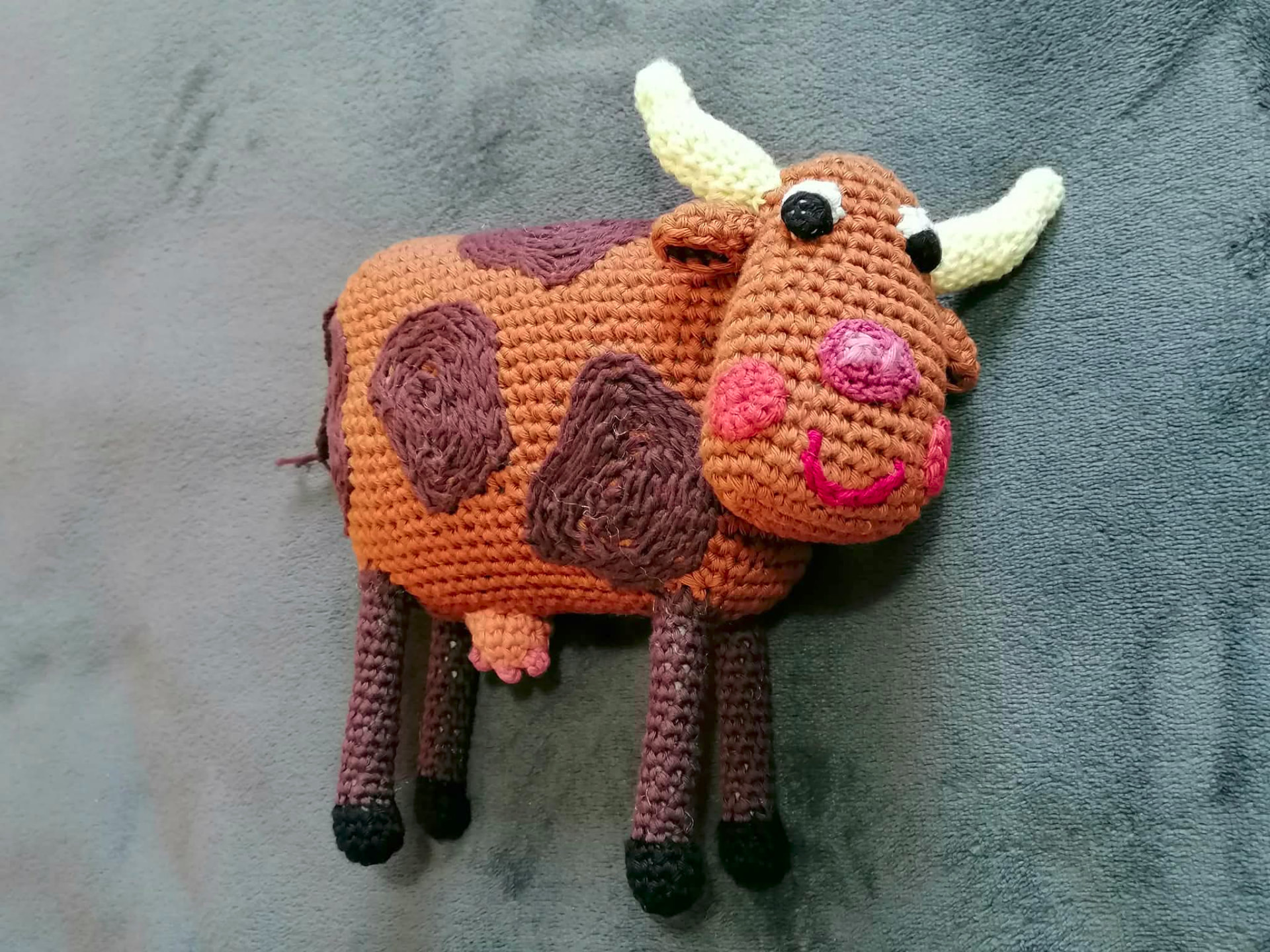 A handmade crocheted cow with a warm brown face, dark spots and cream horns, shown in full