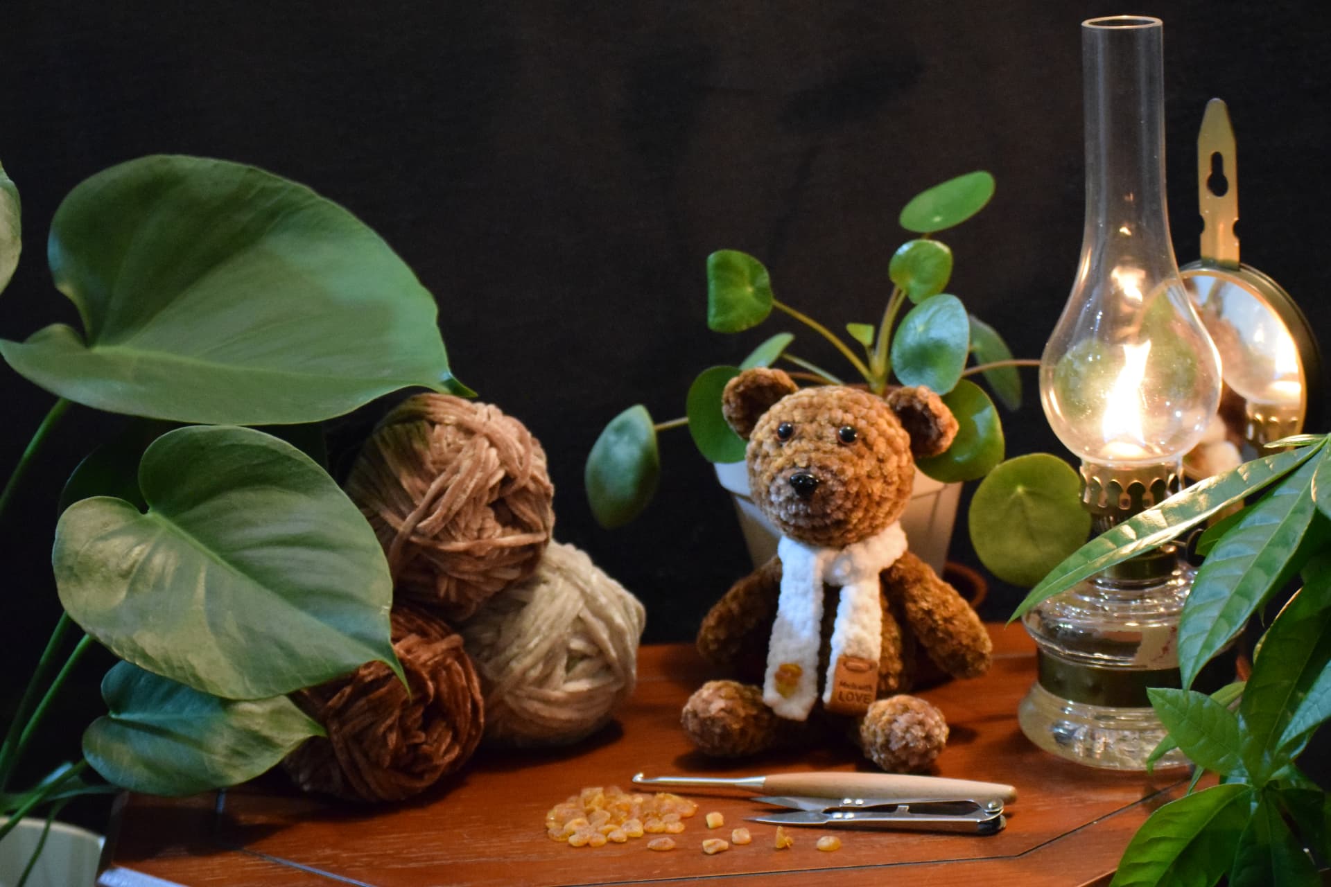 Handmade teddy bear on a wooden table beside chenille yarn, a crochet hook and green plants in warm lamplight